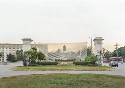 color image of street scene; grass and paved street in foreground with dark gray car on R; small, landscaped area at center with flowers, hedges, palm trees and a large banner hanging between two columns; monument/statue of figure on horse silhouetted against banner; tan building on L in background; moving vehicle on R in background
