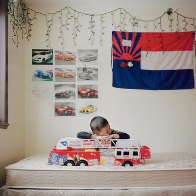 image of a little boy resting his head on his arms on top of a large toy fire engine on a bare mattress; red, white, and blue flag on wall at right with a rising sun in UL and three bold stripes; pictures of sports cars on wall to left; icicle Christmas lights above flag and pictures of cars