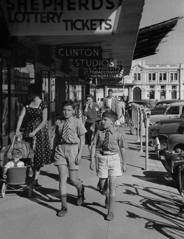 Black and white photograph of two boys wearing ties and shorts walking down a "mainstreet" type sidewalk