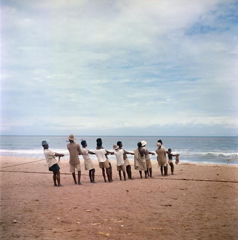 Color photograph of figures standing on a beach pulling a rope