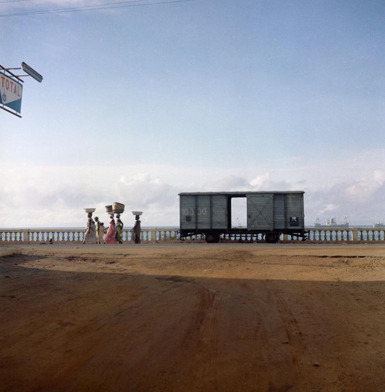 Color image of a train car against a railing with water in the background; figures balancing objects on their heads walk toward the viewer's left